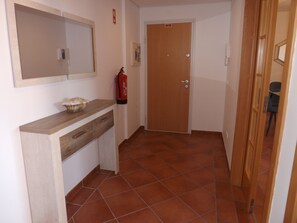 Un couloir avec une porte en bois, un extincteur, un miroir et une petite étagère sur laquelle est posé un objet décoratif.