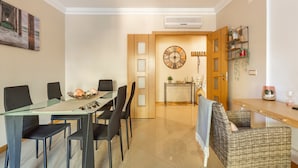 A modern dining area with a glass table, black chairs, a wicker chair, a wooden door, and a wall clock. - Solmar Garden View Apartment: Pool, Wi-Fi & Air Conditioning (Albufeira)