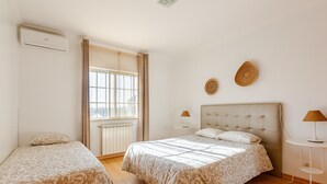 Une chambre à coucher comprenant un lit, deux tables de chevet, une fenêtre avec des rideaux et un appareil de climatisation.