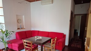Un salon avec un canapé rouge, une table à manger en bois et des chaises. On y trouve également une plante en pot, une horloge au mur et un appareil de climatisation.
