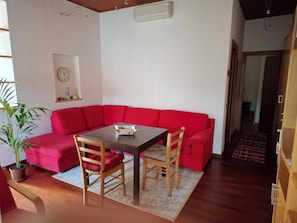 Ein Wohnzimmer mit rotem Sofa, Esstisch aus Holz und Stühlen. Eine Topfpflanze, eine Wanduhr und eine Klimaanlage sind ebenfalls vorhanden.