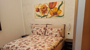Une chambre à coucher avec un lit, une table de chevet, une lampe fixée au mur et un tableau représentant des fleurs.