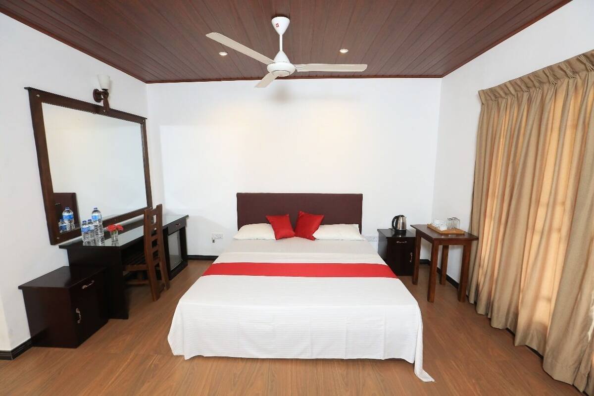Habitación de hotel con cama, escritorio, silla, mesita de noche, espejo, ventilador de techo y ventana con cortinas.