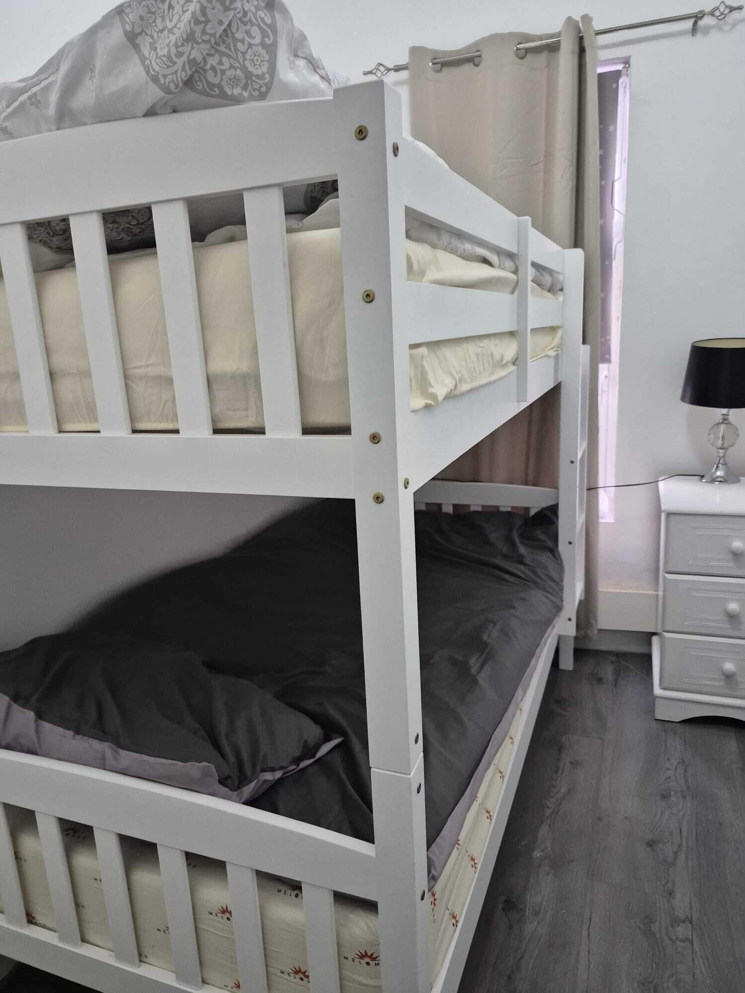 Ein Etagenbett mit einem unterem und einem oberem Bett, ein Nachttisch mit Schubladen und ein Fenster mit Vorhängen.