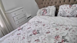 Una cama bien hecha con una colcha de estampado floral y almohadas a juego, un cabecero capitonado y un aparador blanco con cajones.