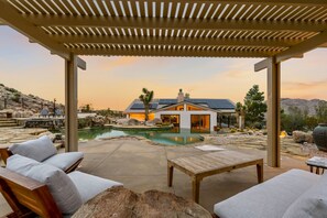 A covered outdoor area with a pool, lounge chairs, and a view of a house in the background. - Breakaway By Homestead Modern (Joshua Tree)