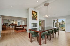 A spacious living area with a dining table, green upholstered chairs, a kitchen in the background, and a large window offering a view of cacti. - Breakaway By Homestead Modern (Joshua Tree)