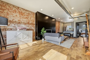 A modern living room with a brick wall, white cabinets, a grey sofa, and a dining area.