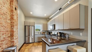 A modern kitchen with stainless steel appliances, a brick wall, and a large island with bar stools. - The Modern Loft at the foot of the Bighorns (Buffalo)