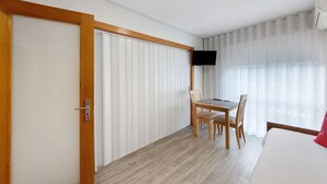 Una camera d'albergo con una porta in legno, un letto con cuscino rosso, un tavolo con sedie, una TV a parete e una finestra con tende leggere.
