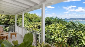 A covered outdoor area with a white railing, a wooden bench, and lush greenery. - Villa OSUN (Les Terres Basses)