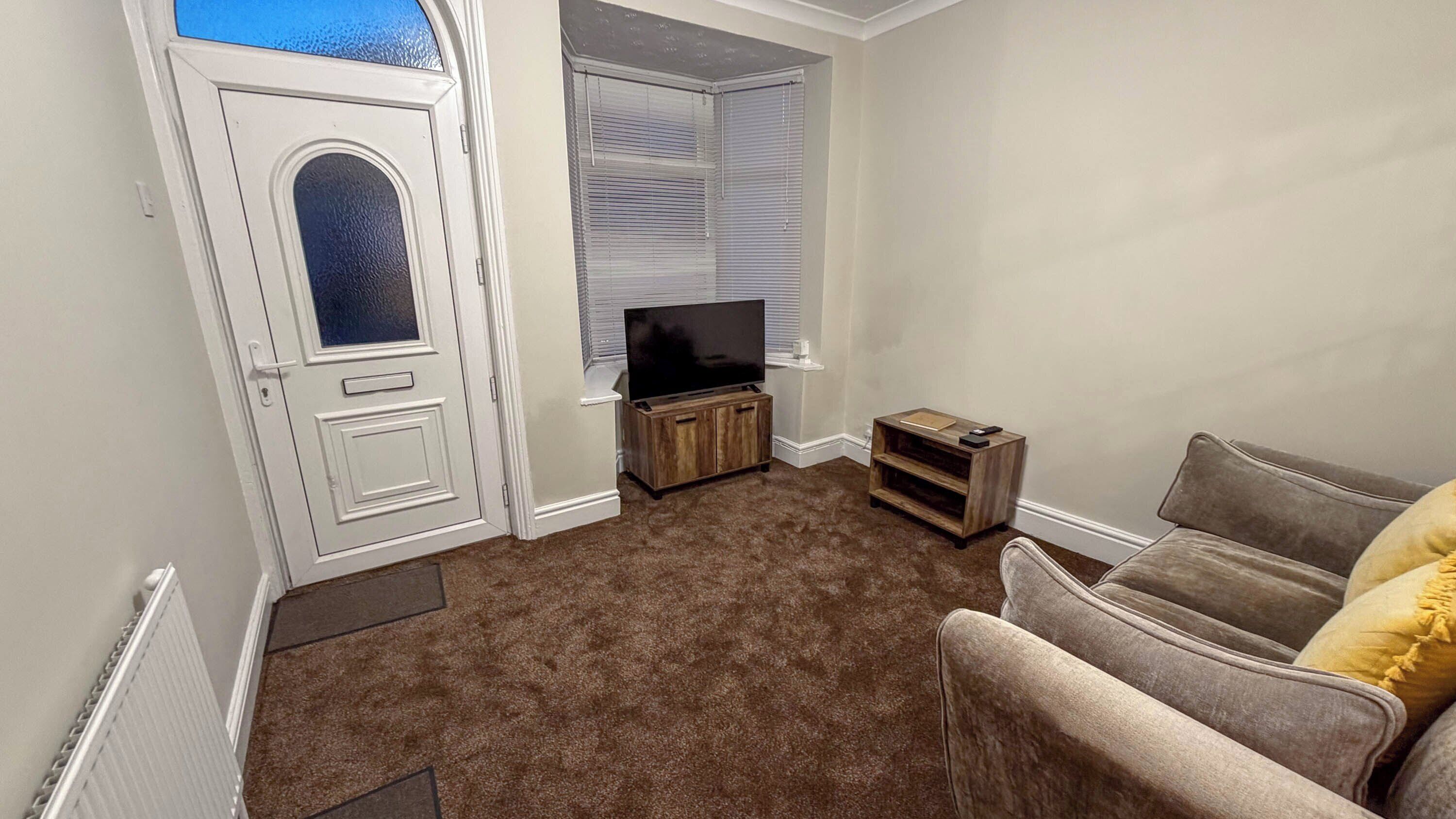 A living room with a brown carpet, a grey sofa, a television on a wooden stand, and a white door with a glass panel.