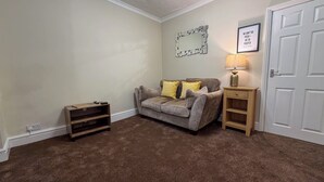A living room with a grey sofa, a wooden side table with a lamp, a small wooden shelf, and a white door. - Family/business - Parking - Wifi - Sleeps 3 (Barnsley)