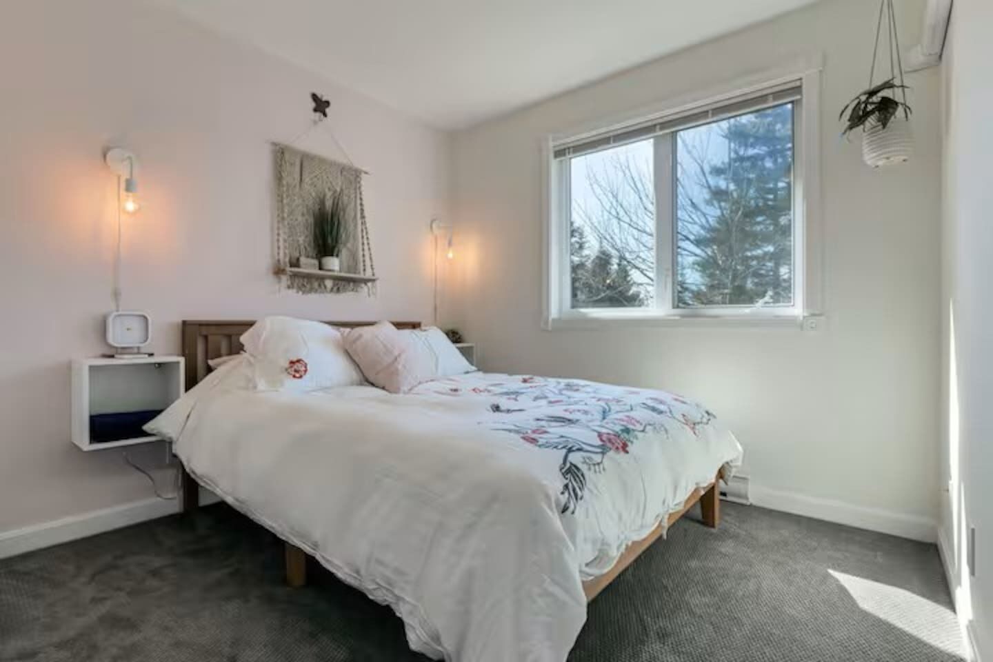 Ein Schlafzimmer mit einem Bett, Nachttischen, einem an der Wand befestigten Regal und einem Fenster mit Blick auf Bäume.