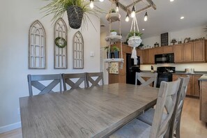 A dining area with a wooden table and chairs, a kitchen with wooden cabinets and appliances, and decorative plants. - Entire Pine Ridge - 2 Attached Four Bedroom Condos (Bar Harbor)