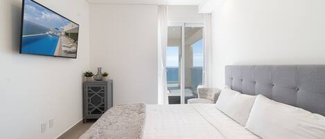 A modern bedroom with a large bed, a television mounted on the wall, and a view of the ocean through a sliding glass door.
