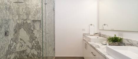 Un baño moderno con ducha envuelta en vidrio, paredes de mármol, un espejo grande y una encimera de mármol con lavabo y tocador.