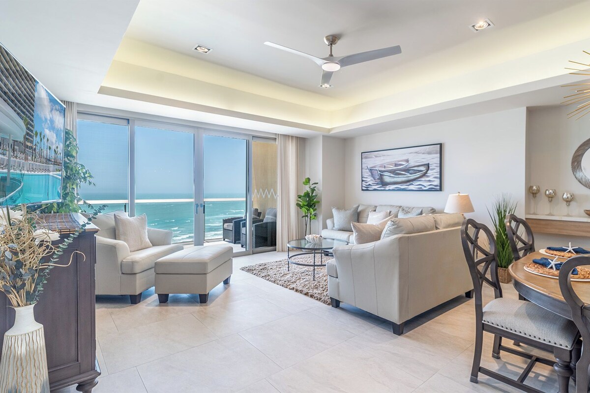 A modern living room with a large window overlooking the ocean, a ceiling fan, and a dining area with a round table.