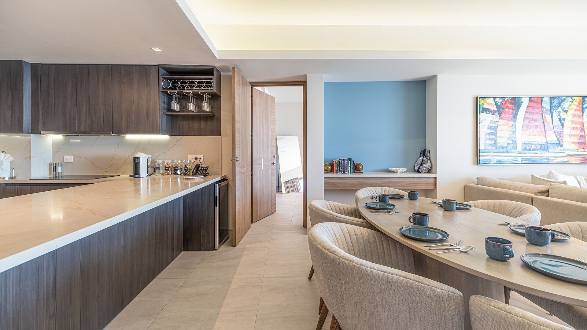 Une cuisine moderne avec des meubles de couleur bois foncé, un plan de travail en marbre et un coin repas comprenant une table et des chaises aux couleurs claires.