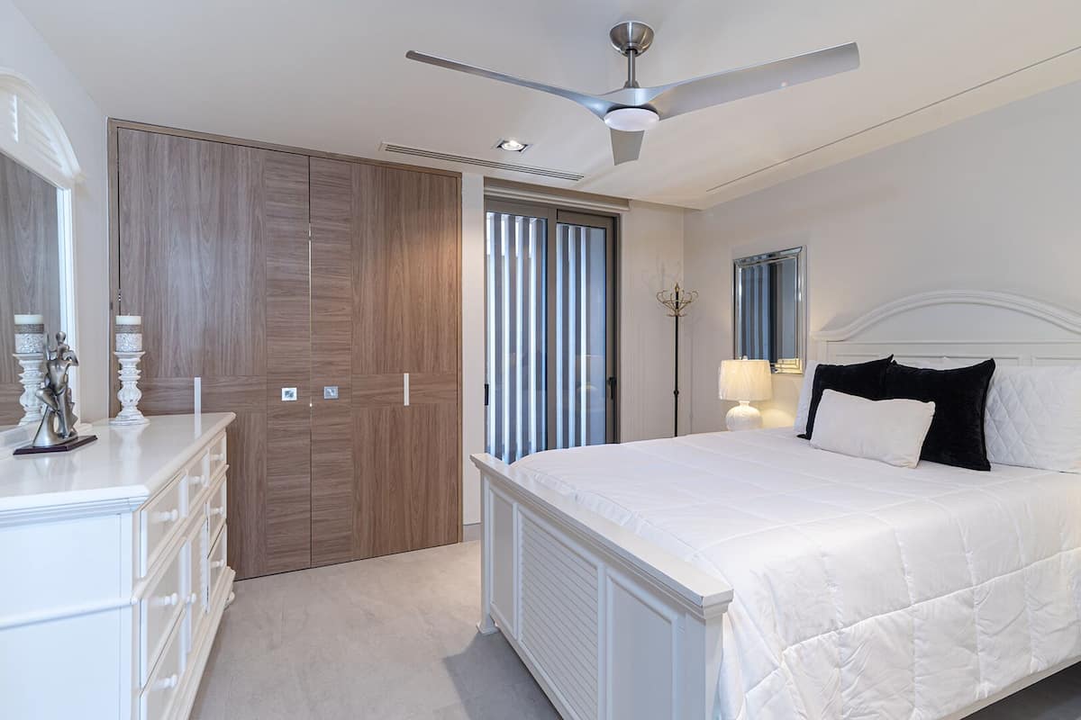 Une chambre à coucher comprenant un lit, des tables de chevet, une commode, un ventilateur de plafond et une grande fenêtre avec des stores.