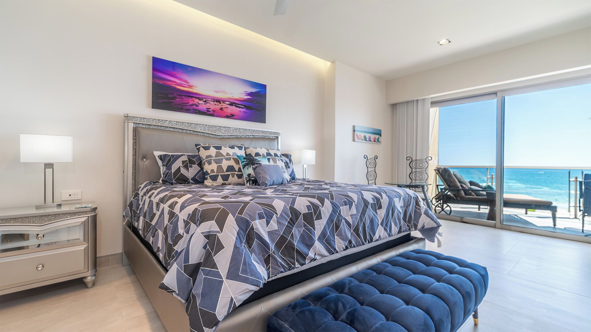 A modern bedroom with a large bed, a nightstand, a bench, and a view of the ocean.