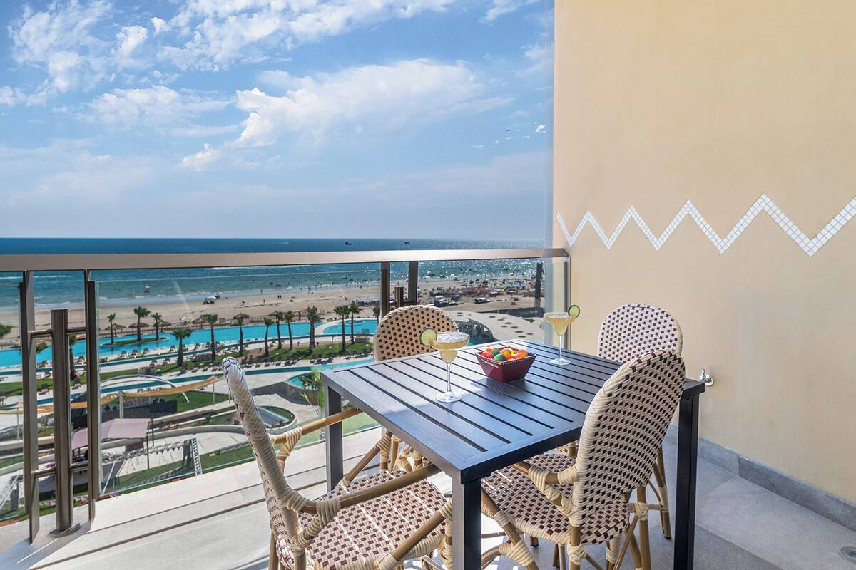 Un balcon avec des fauteuils en osier, une table et une vue sur la plage et la piscine.