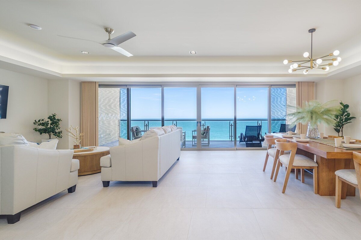 Un salon spacieux avec un grand canapé, un coin repas avec des chaises en bois et une vue sur l’océan grâce à de grandes portes vitrées.