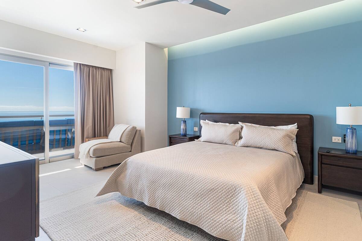 Une chambre d’hôtel moderne avec un grand lit, deux tables de chevet avec des lampes, un canapé et une vue sur l’océan.