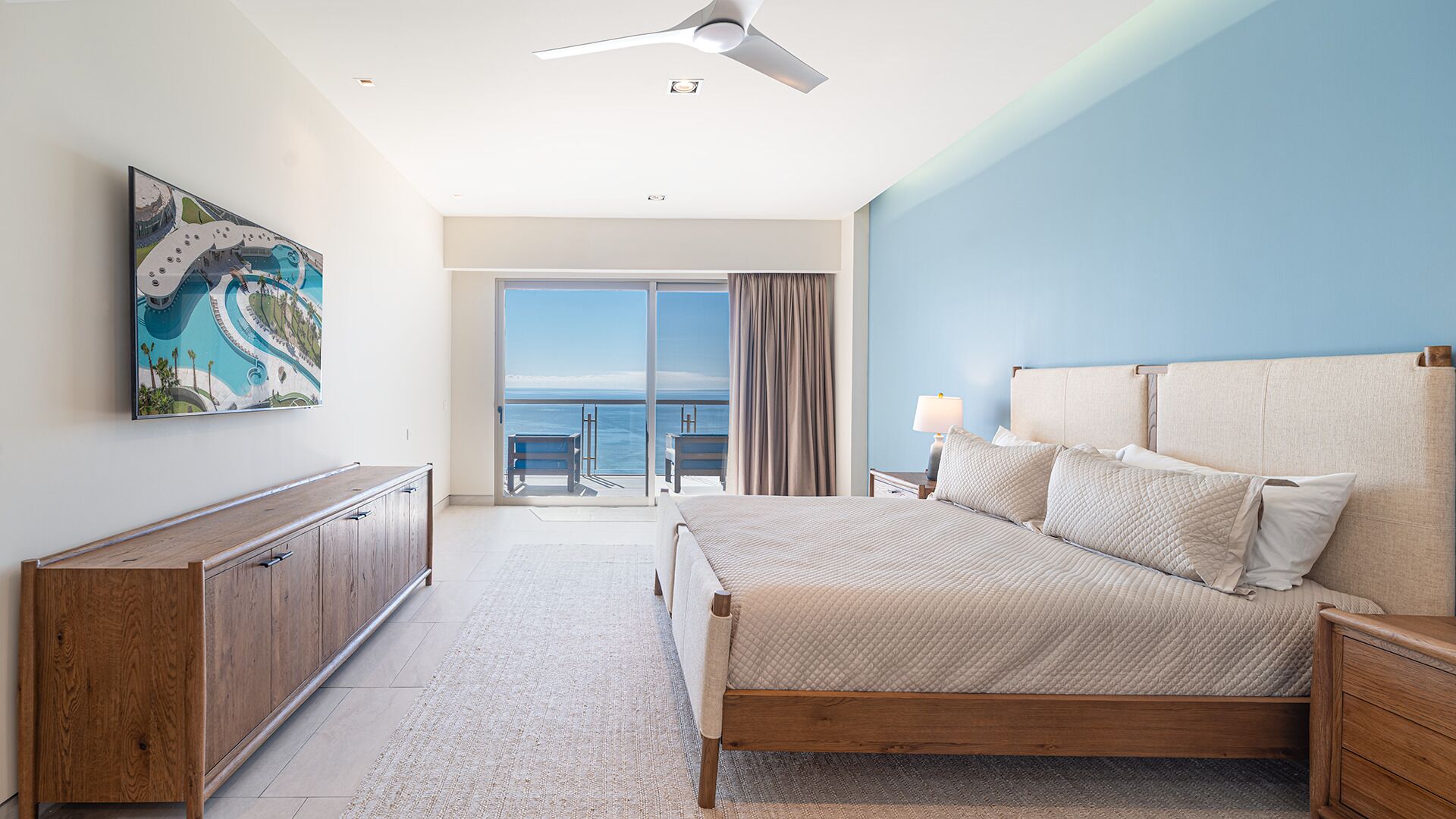 A bedroom with a bed, a nightstand, a wooden dresser, a ceiling fan, and a large window with a view of the ocean.