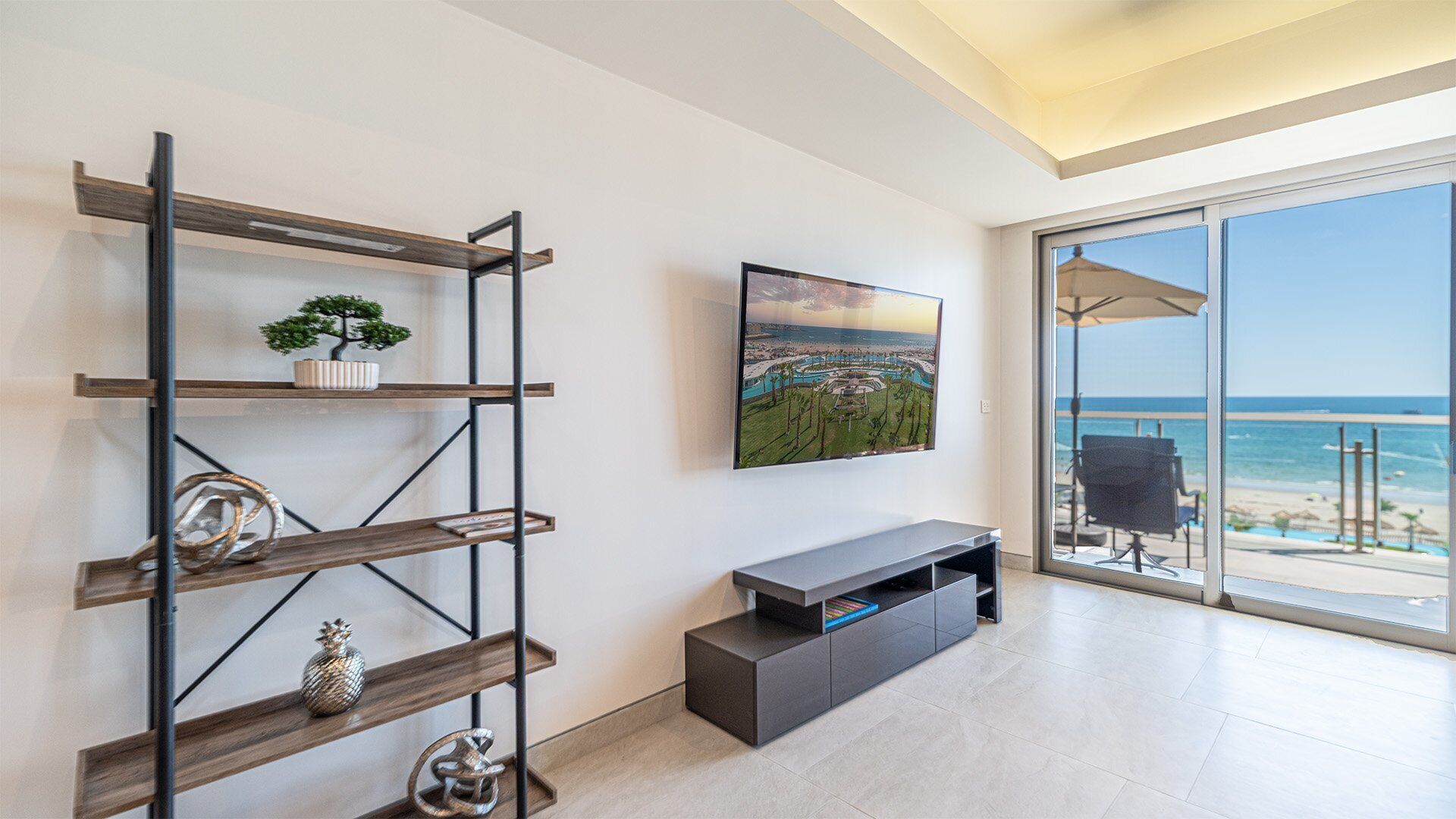 A modern living room with a large window overlooking a beach, a mounted TV, and a minimalist shelving unit.