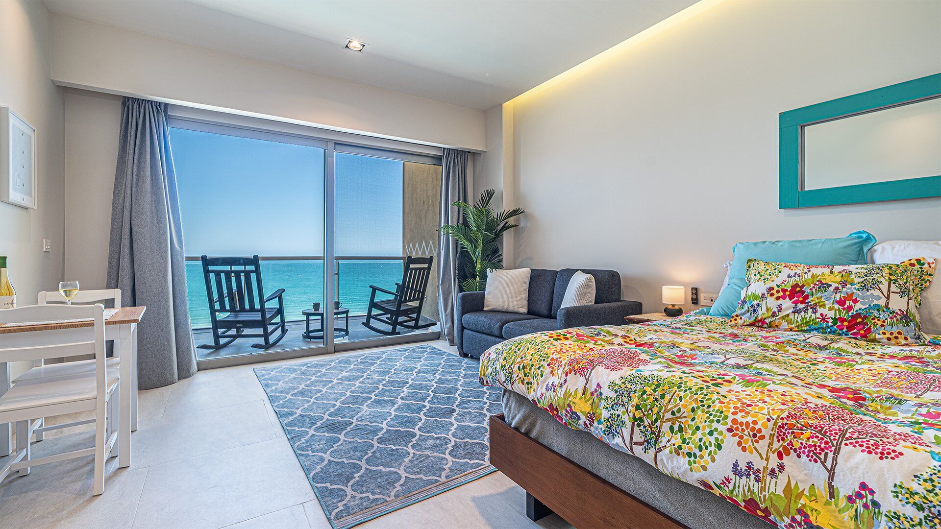 Una habitación de hotel con balcón, una cama con colcha floral de colores, un sofá gris y una zona de comedor con mesa y sillas.