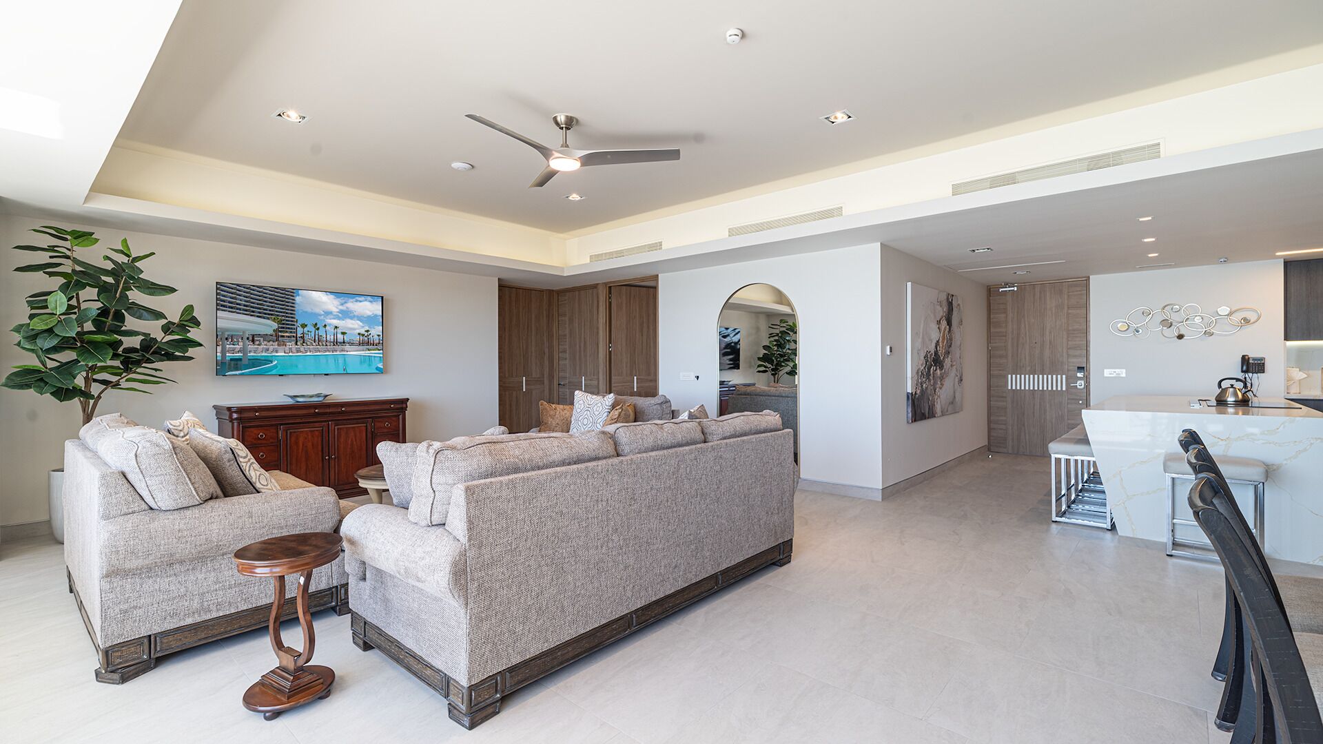 A spacious living area with a large sofa, a coffee table, and a TV mounted on the wall. There is a plant, a wooden cabinet, and a kitchen area visible in the background.