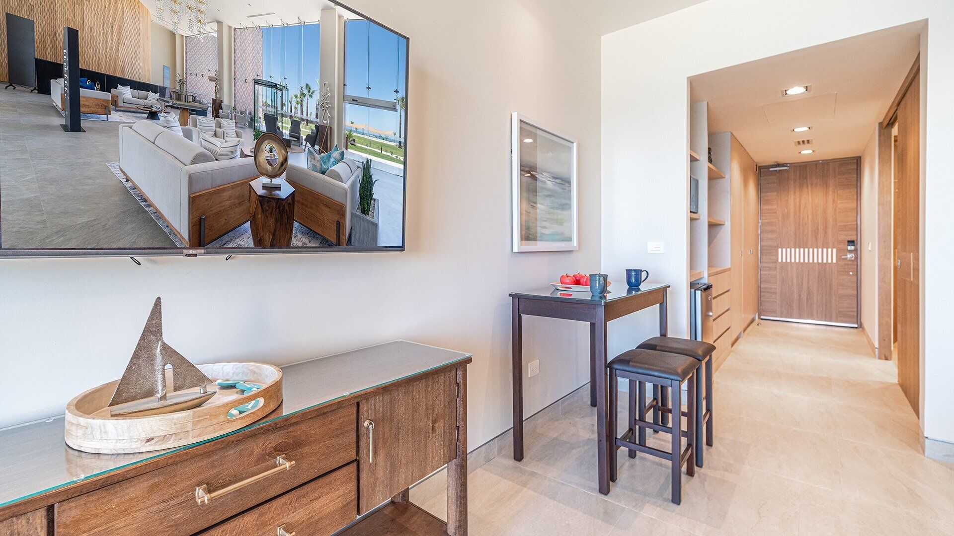 Un hall d’hôtel moderne avec un grand miroir qui reflète la pièce, un bureau en bois doté de tiroirs, une petite table avec une chaise, et une vue sur la plage grâce à de grandes fenêtres.