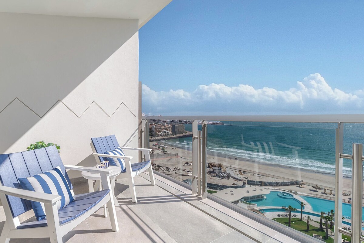 Un balcón con dos sillones reclinables a rayas azules, una mesita y vistas a la playa y al océano.