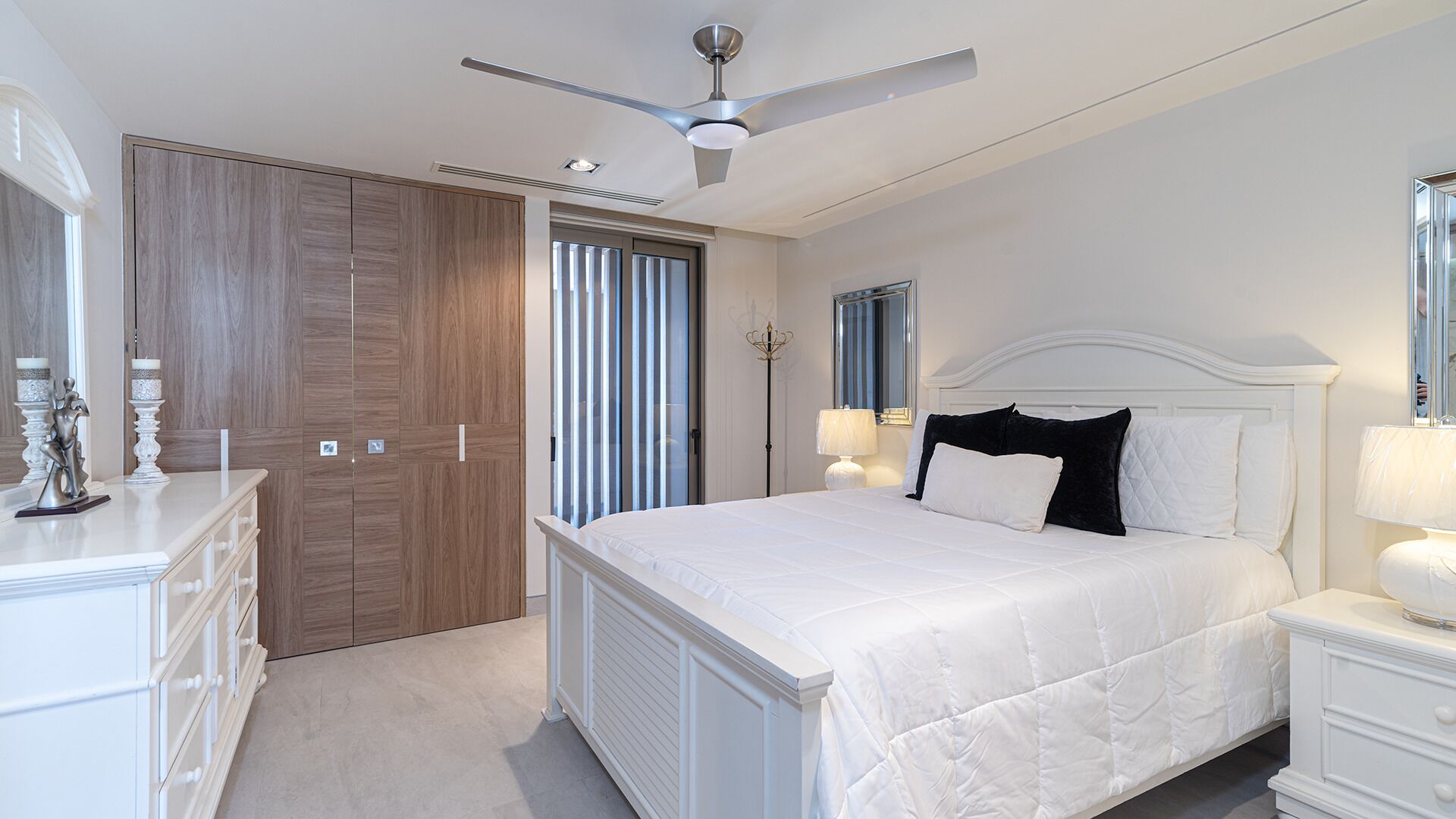 Ein Schlafzimmer mit Bett, Nachttischen, einem Schminktisch, einem Deckenventilator und einem großen Fenster mit Jalousien.