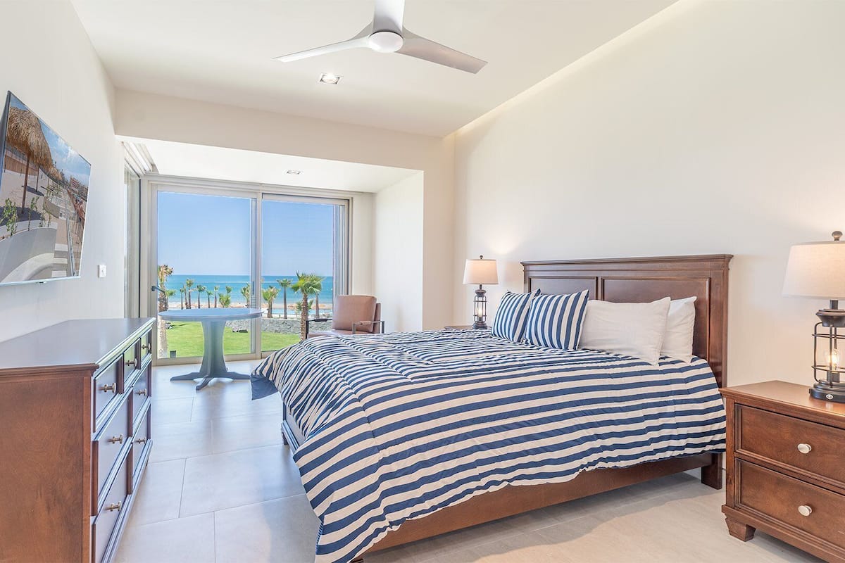 Ein Schlafzimmer mit einem großen Bett, einem Nachttisch, einem Deckenventilator, einem Fenster mit Blick auf den Strand und einem Tisch mit Stühlen draußen.
