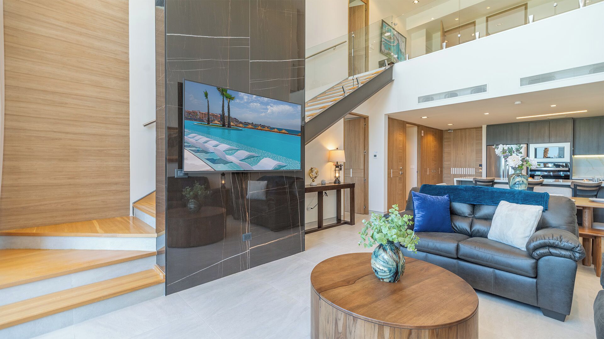 A spacious living area with a modern design, featuring a large flat-screen TV, a comfortable sofa, a wooden coffee table, and a staircase leading to an upper level.