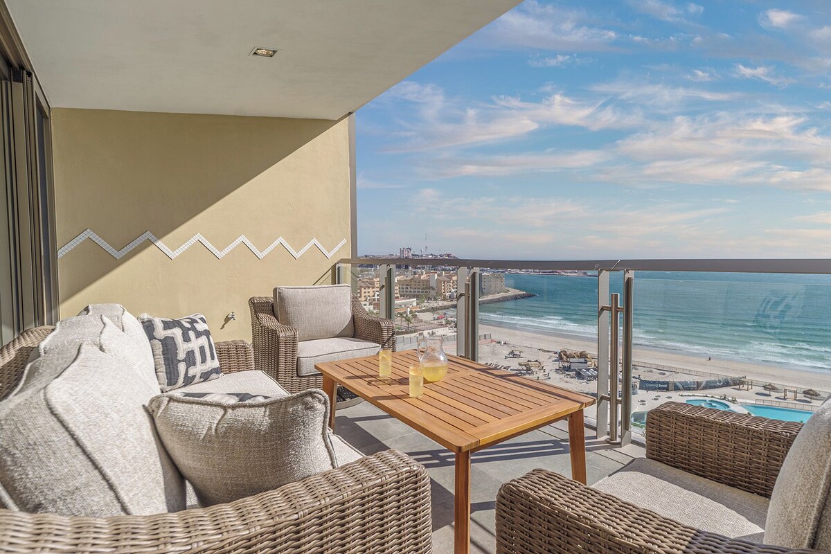 Een balkon met rieten meubilair, een houten tafel en uitzicht op het strand en de zee.