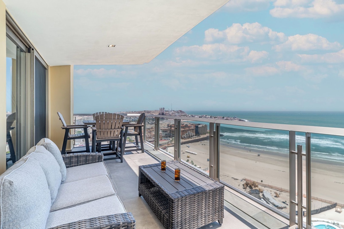 Un balcon doté de meubles en osier, d’une rambarde en verre et offrant une vue sur la plage et l’océan.