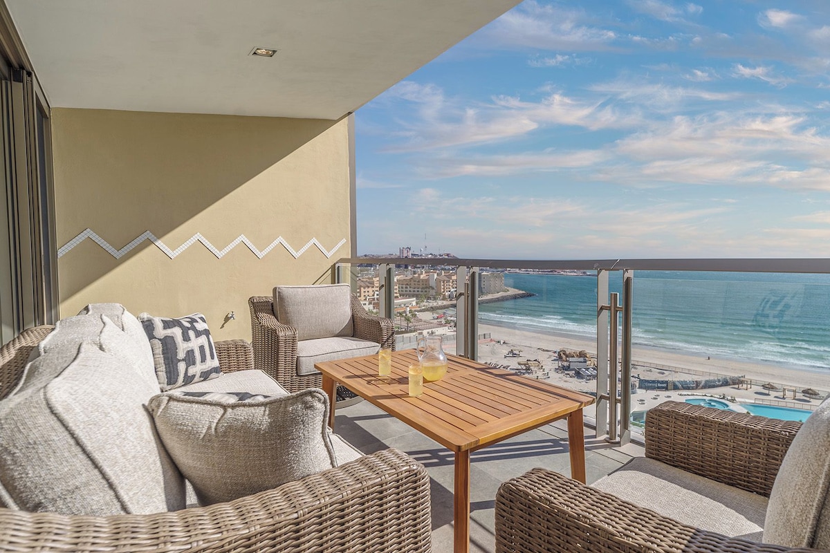 Ein Balkon mit Korbmöbeln, einem Holztisch und Blick auf Strand und Meer.