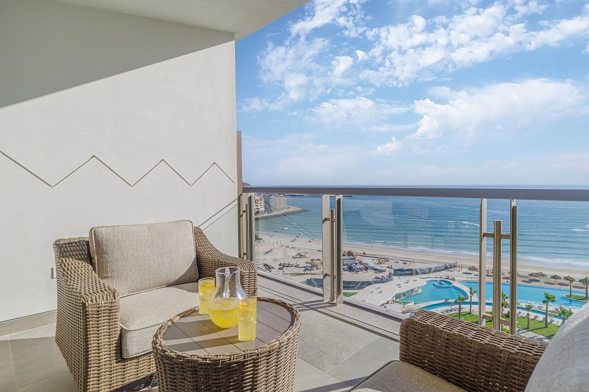 A balcony with wicker furniture, a glass table with drinks, and a view of the beach and ocean.