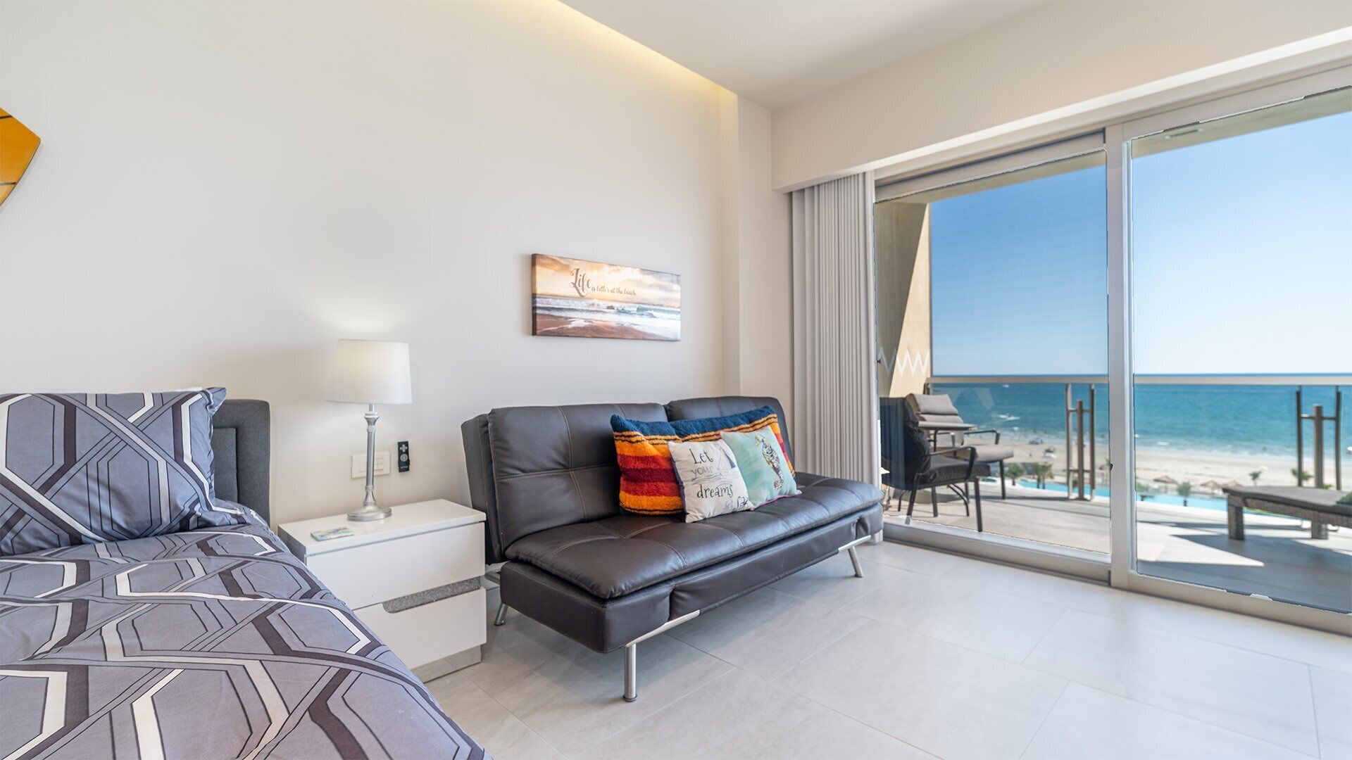 Een moderne hotelkamer met een bed, een bank, een nachtkastje en een ruim balkon met uitzicht op het strand.