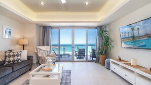Un soggiorno moderno con un'ampia finestra vista mare, un divano confortevole, un tavolino da caffè e un televisore che mostra una spiaggia.