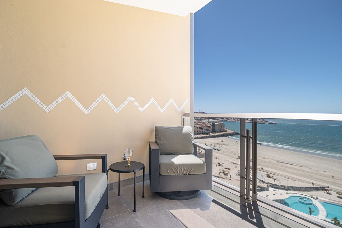 Ein Balkon mit zwei Stühlen, einem kleinen Tisch und einem Glas Wasser. Blick auf einen Strand und ein Schwimmbecken.