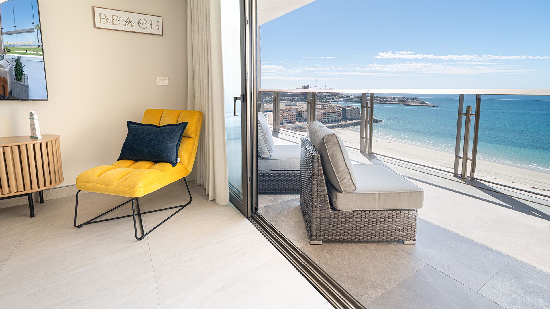 A balcony with a yellow chair and a grey sofa, overlooking a beach and cityscape.