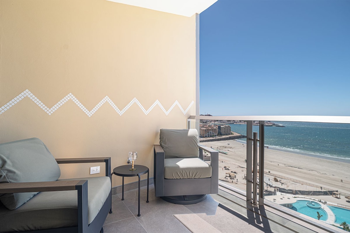 A balcony with two chairs, a small table, and a glass of water. The balcony overlooks a beach and a pool.