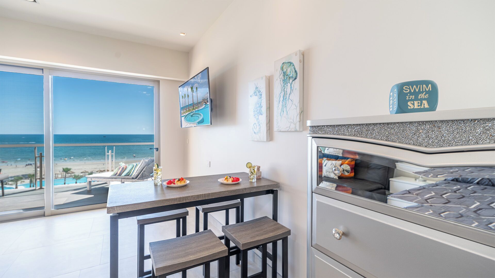 Una habitación de hotel moderna con un balcón grande con vistas a la playa, un área de comedor con mesa y sillas, y obras de arte en las paredes.