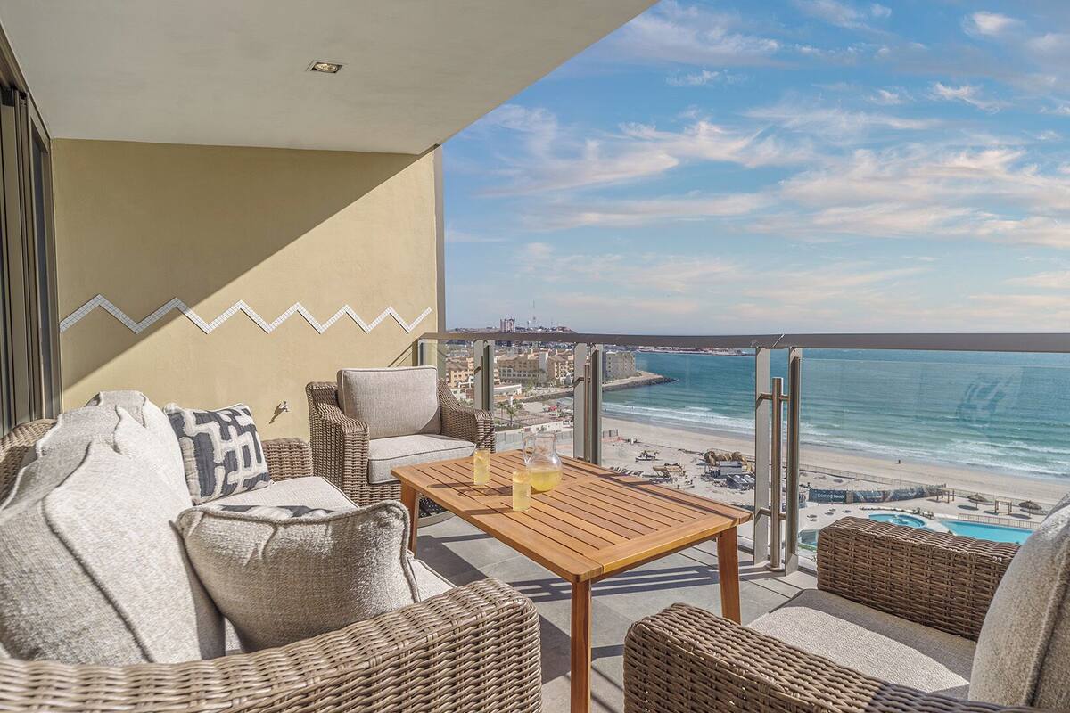 Ein Balkon mit Korbmöbeln, einem Holztisch und Blick auf Strand und Meer.