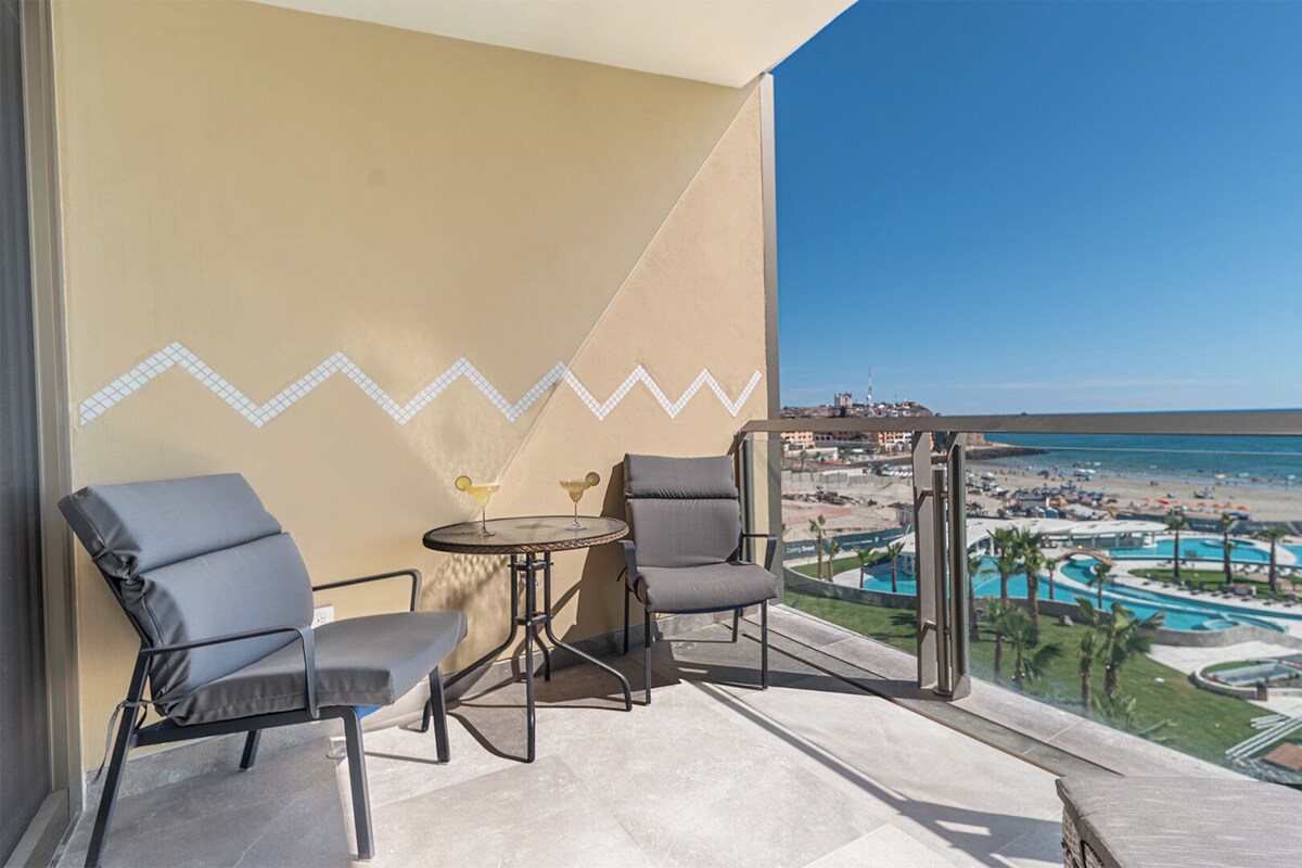 A balcony with two chairs, a small round table, and a view of a beach resort with a pool and palm trees.