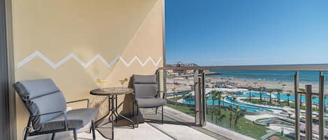 A balcony with two chairs, a small round table, and a view of a beach resort with a pool and palm trees.